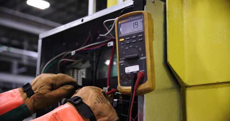 Gloved hands using a multimeter beside an electrical panel